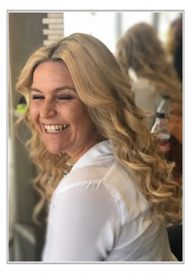 Eine lächelnde blonde Frau in einem weißen Oberteil dreht sich in einem sanft beleuchteten Salon zu einem Spiegel um.
