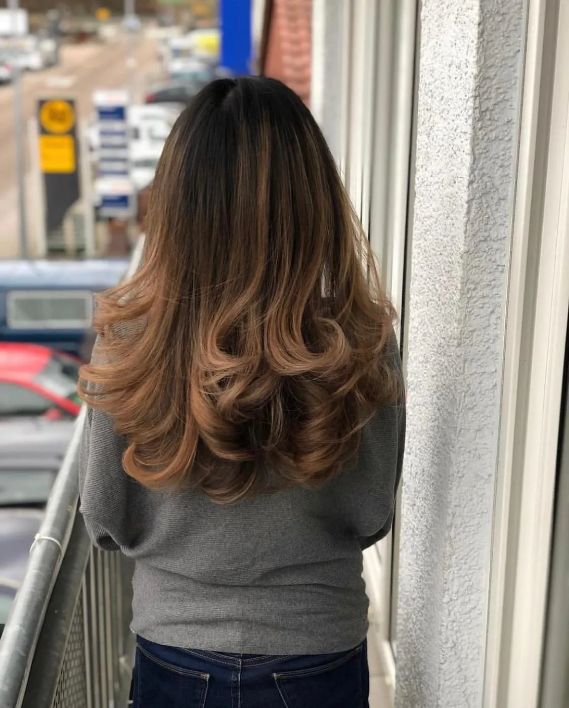 Person mit langen Haaren in einem grauen Pullover, von hinten auf einem Balkon mit Blick auf einen Parkplatz gesehen.