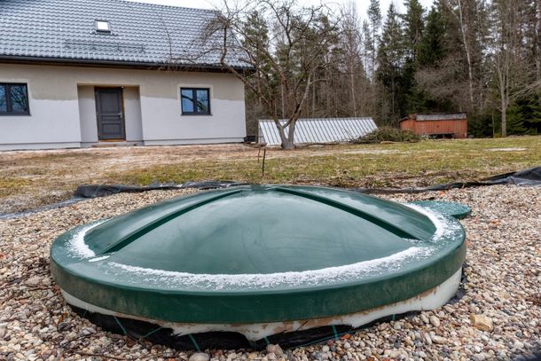 Green round underground tank cover in a gravel yard beside a house with trees in the background