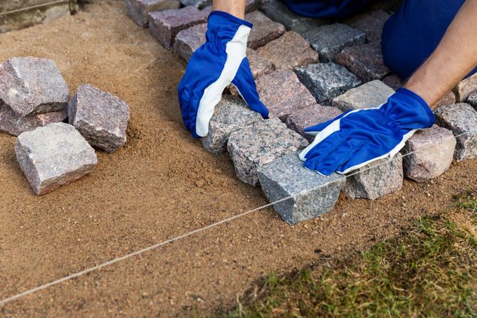 Gloved hands laying gray paving stones along a taut guide string on sand and gravel