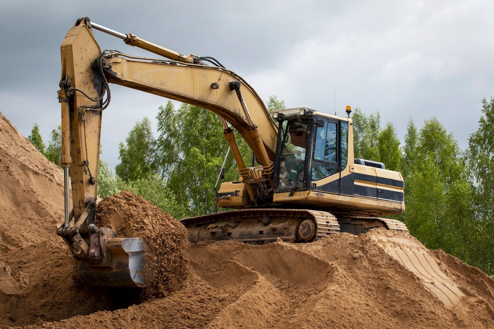 Yellow excavator digging a sand pile at a construction site with trees in the background