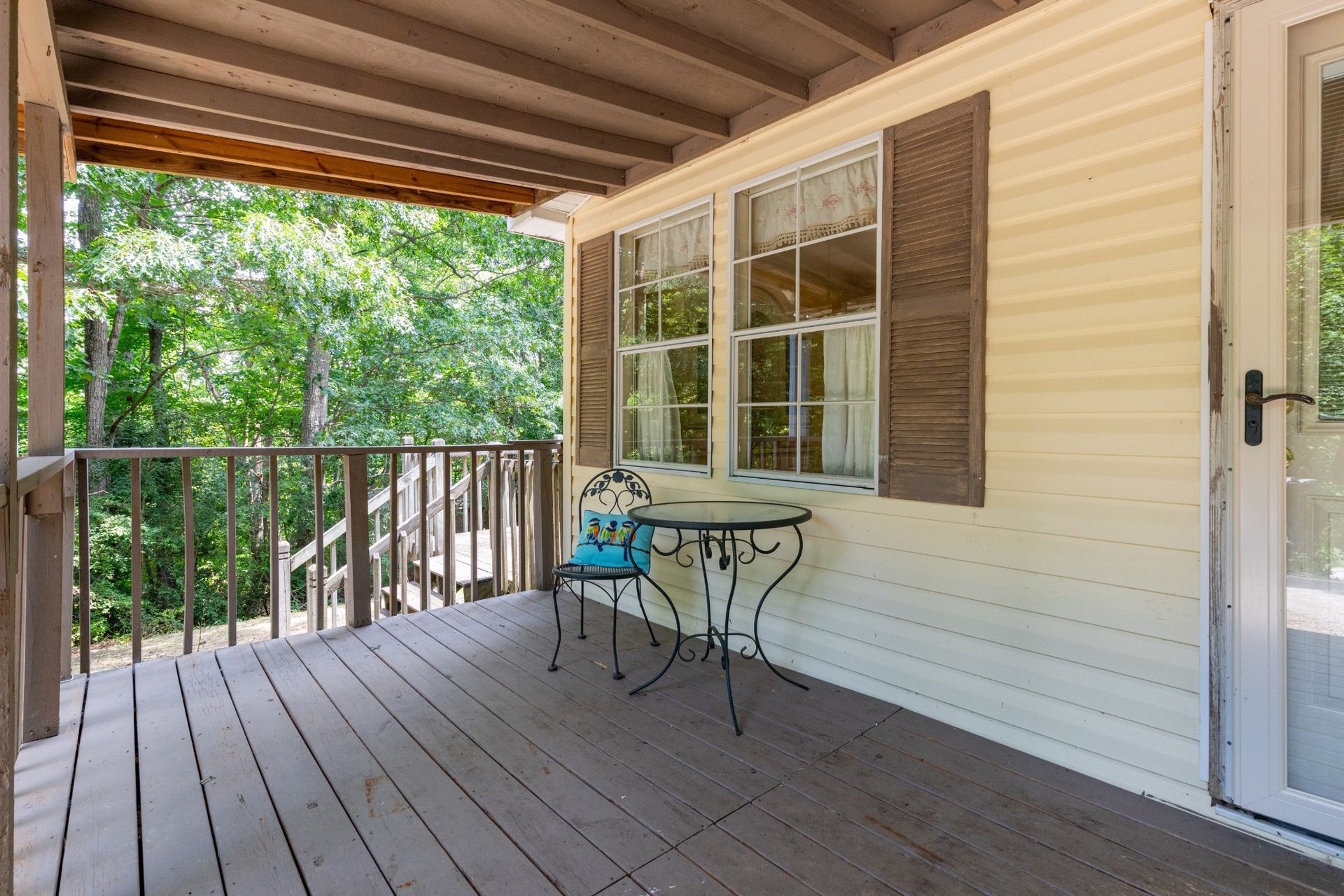 Covered wooden porch with railing, white siding, windows, and a small metal table and chair set.