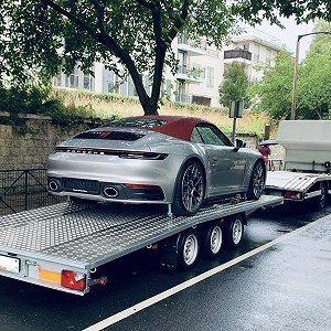 Zilveren Porsche cabriolet op een aanhanger, met rode kap, geparkeerd op een natte weg.