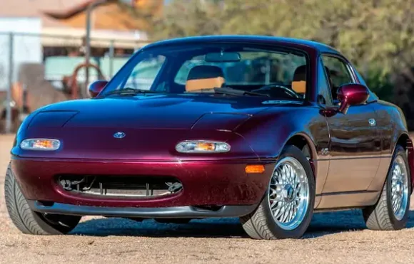 Bordeauxrode Mazda Miata sportwagen met hardtop, chromen wielen, geparkeerd in de buitenlucht.