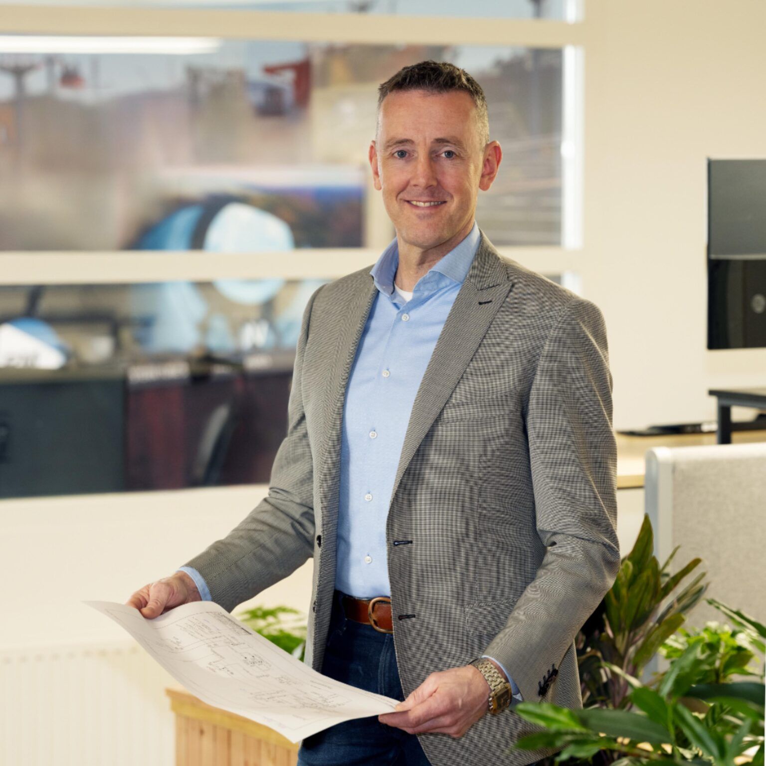 Man in blazer met architectuurtekeningen, lachend in een kantooromgeving met planten en een raam.