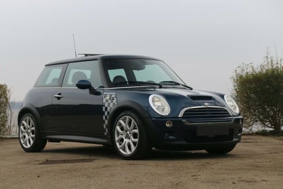 Donkerblauwe Mini Cooper geparkeerd op een onverharde ondergrond met een waterige achtergrond. Dambordmotief op de voorspatbord.
