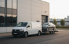 Witte bestelwagen met een auto op een aanhanger voor een modern industrieel gebouw.