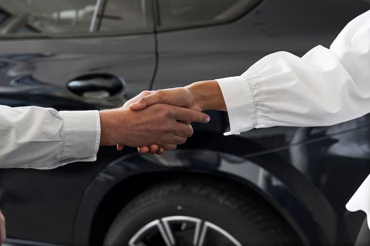 Twee mensen schudden elkaar de hand voor een donkere auto, mogelijk een autodealer.