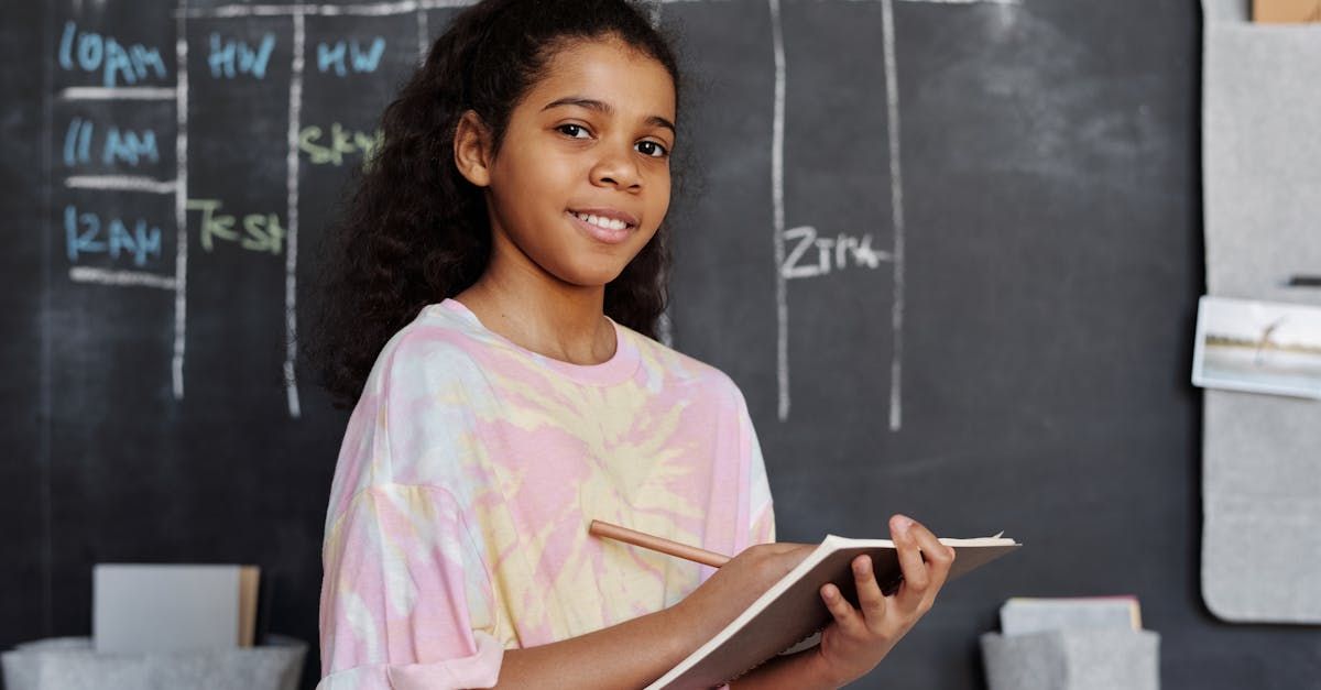 A young girl is standing in front of a blackboard holding a notebook and a pen.