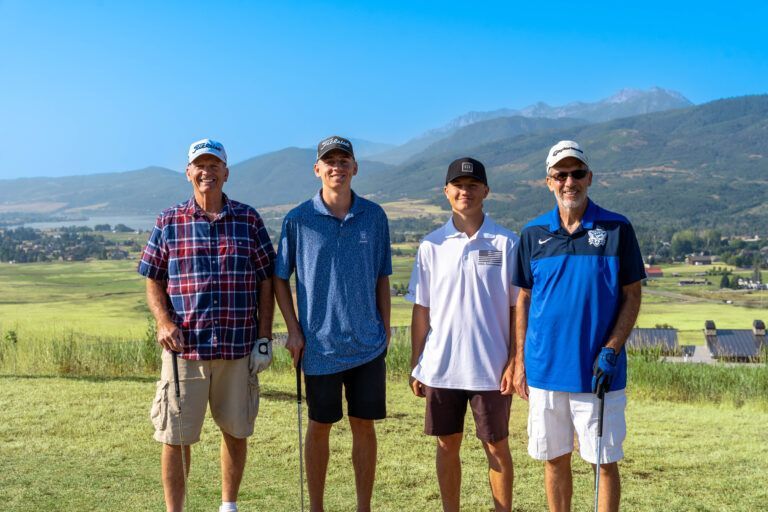 Four golfers stand smiling in a line on a grassy hill overlooking a valley and mountain range under a clear blue sky.
