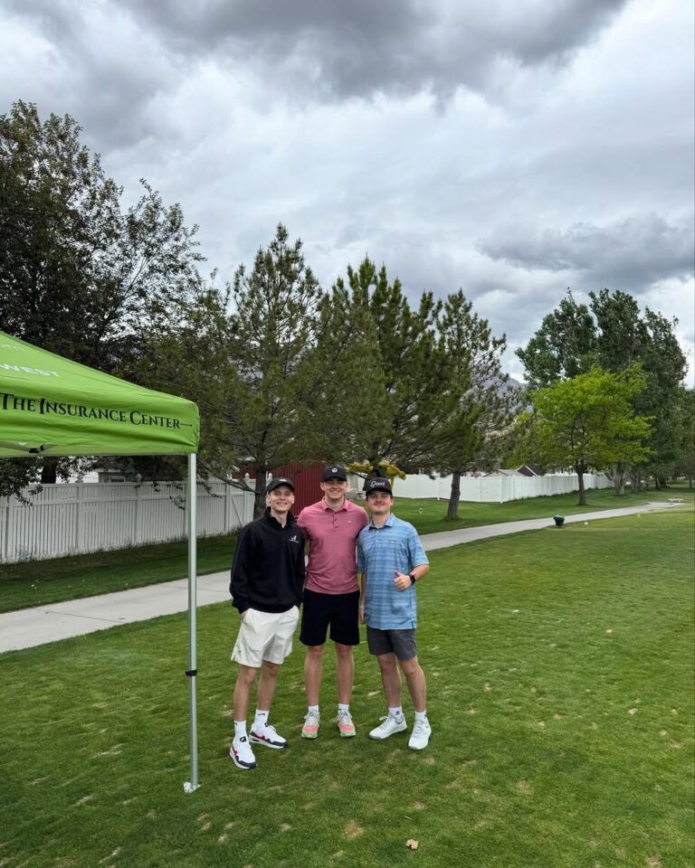 Three people stand smiling on a grassy golf course under a cloudy sky next to a green tent.