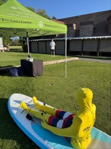 A yellow-clad mannequin sits on a paddleboard on a grassy field near an insurance booth and a standing person.