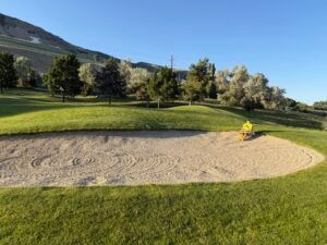 A sand bunker on a golf course with a yellow rake resting on the green grass edge against a backdrop of trees and a hill.
