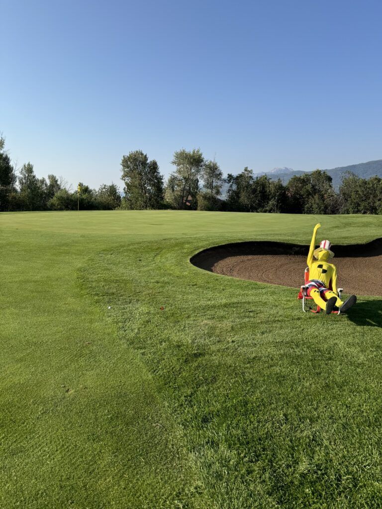 A yellow crash test dummy sits on a red chair at the edge of a golf course sand bunker under a clear blue sky.