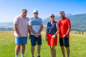 Four golfers wearing hats and sunglasses stand on a sunny course with mountains in the background.