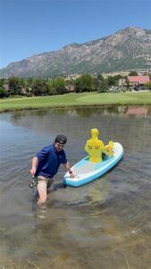 A person in a blue shirt standing in a lake, holding a light blue paddleboard with a yellow mannequin sitting on it.