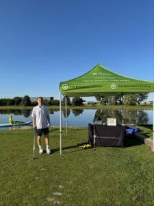 A person stands beside a green tent next to a lake on a sunny day.