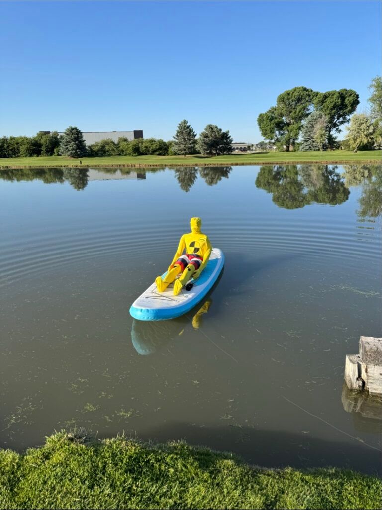 A yellow crash test dummy sits on a light blue and white stand-up paddleboard on a calm, reflective pond.