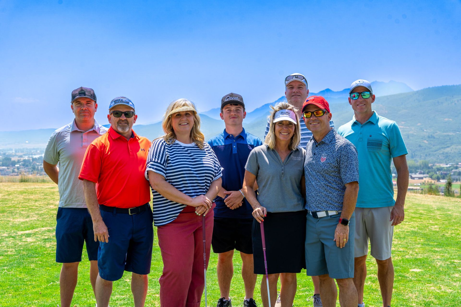 A group of eight people stand in a line on a grassy hill outdoors with mountains in the background under a blue sky.