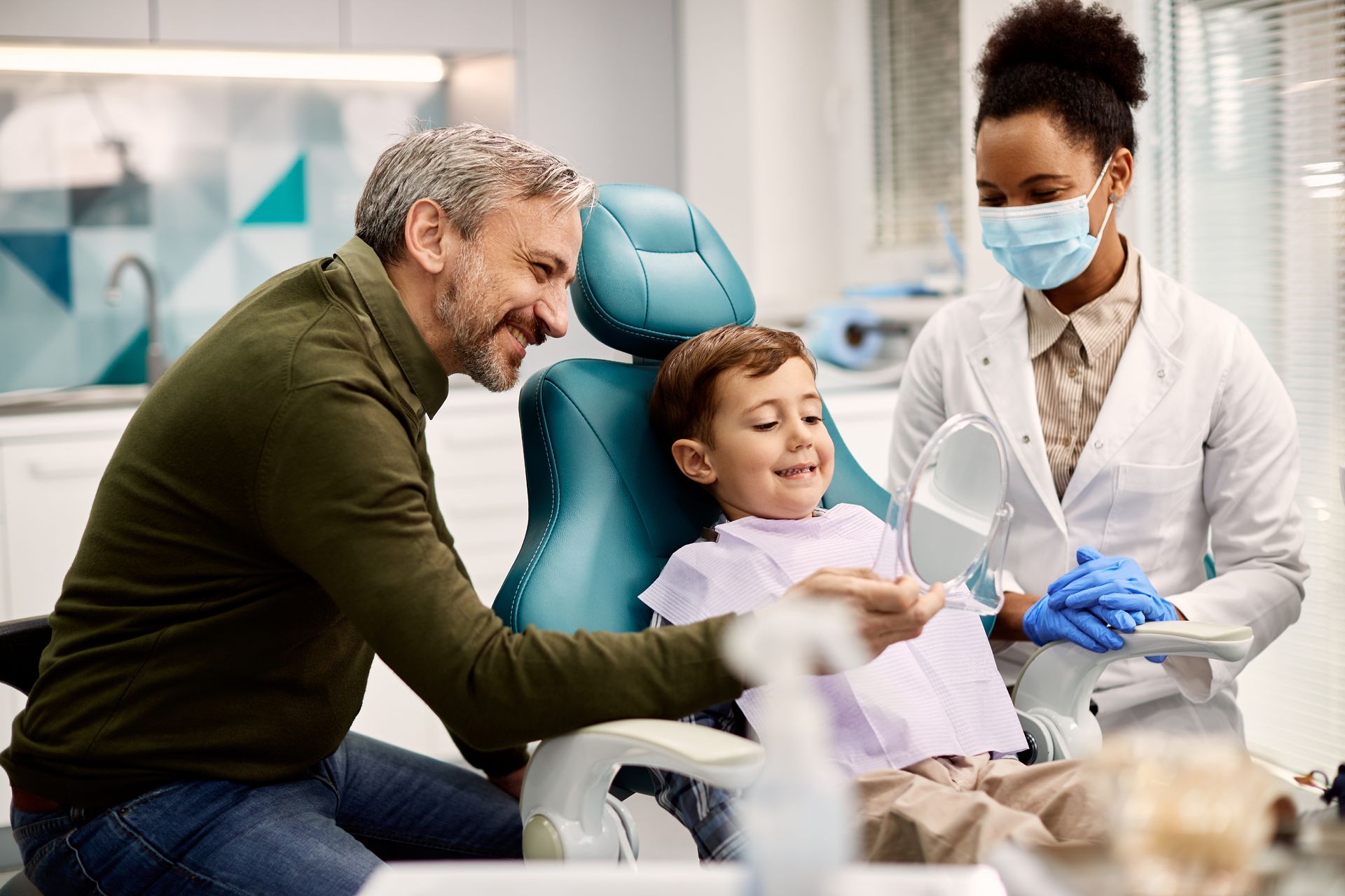 boy looking at his teeth in a mirror while being with his father at dentist's office.
