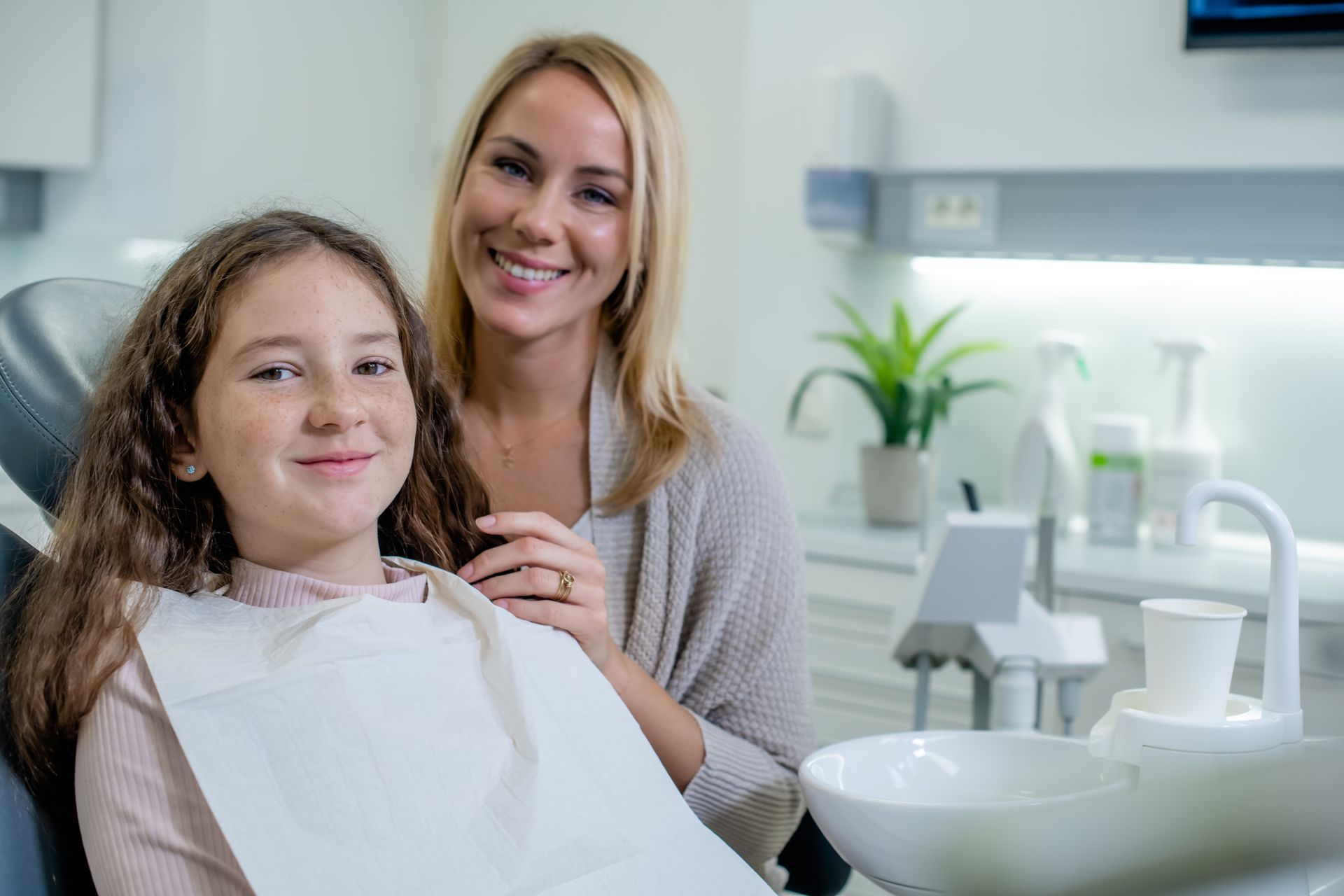 Young girl smiling in a dental chair next to her mother during a checkup with their family dentist.