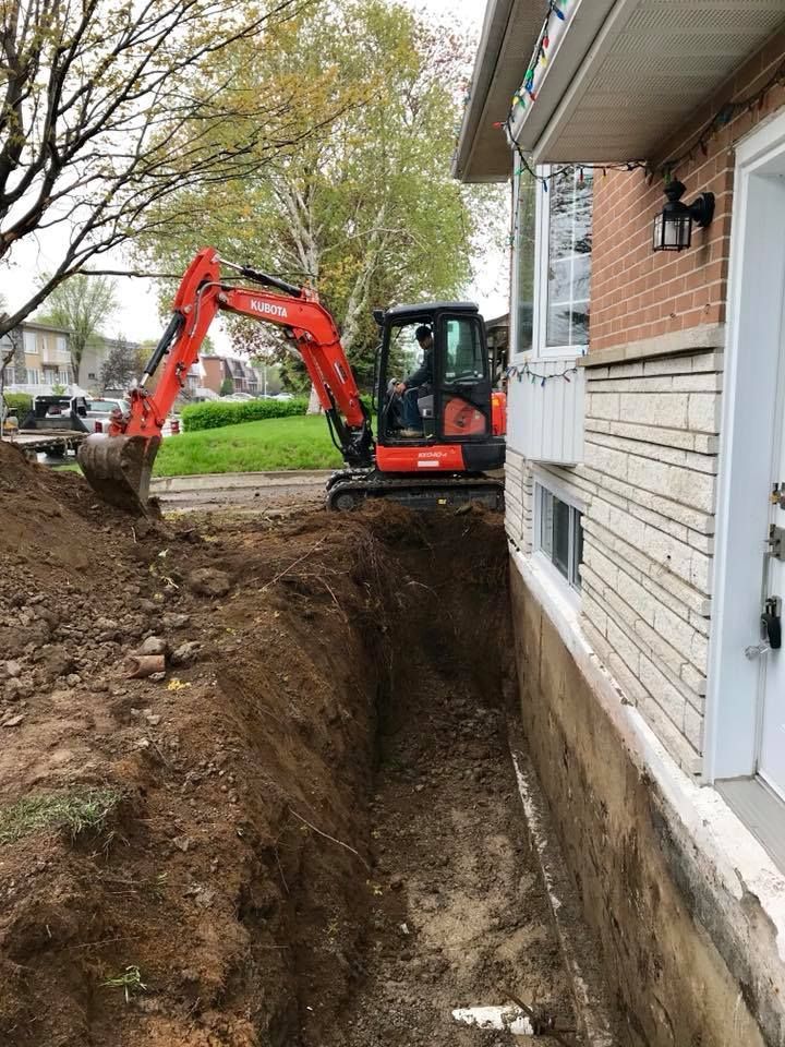 Une pelleteuse orange creuse une profonde tranchée le long des fondations en briques d'une maison d'habitation.