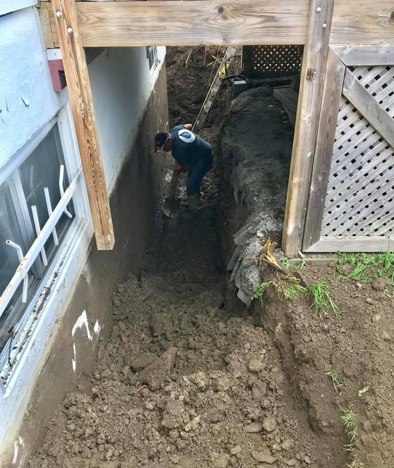 Une personne vêtue d'une chemise sombre travaille dans une tranchée profonde et étroite creusée le long des fondations d'une maison, sous une terrasse en bois.