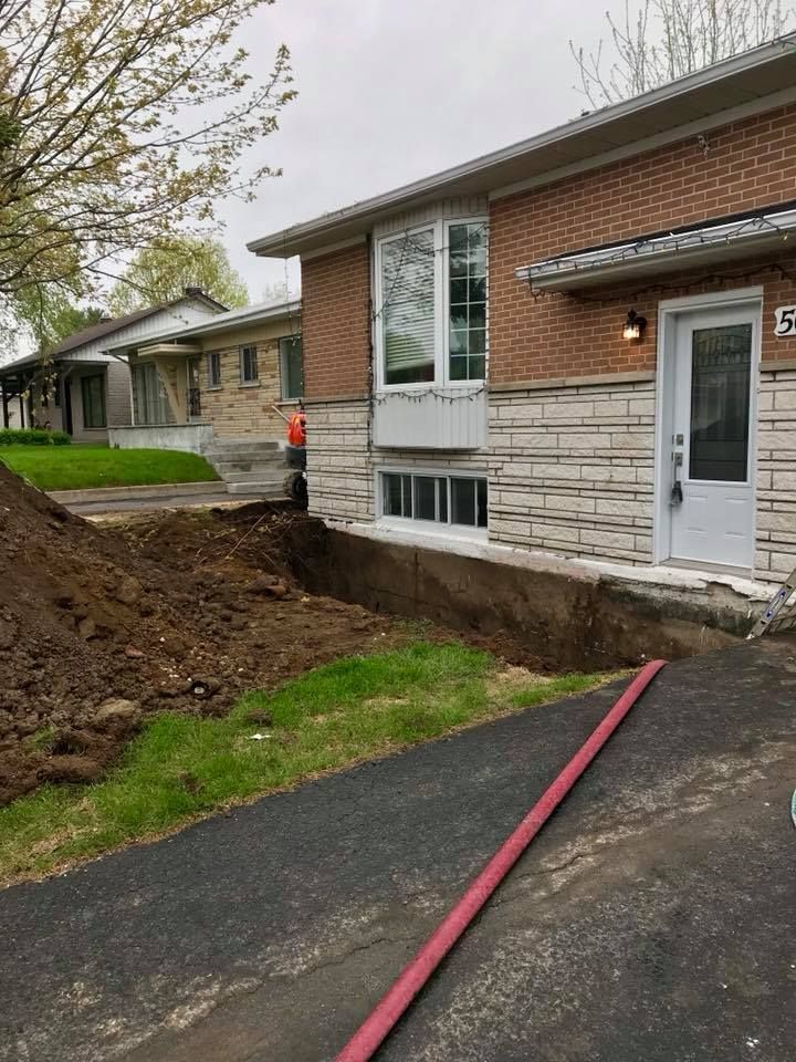 L'extérieur d'une maison présente une large tranchée creusée le long du mur de façade, à côté d'une allée pavée, et un tuyau d'arrosage rouge au sol.