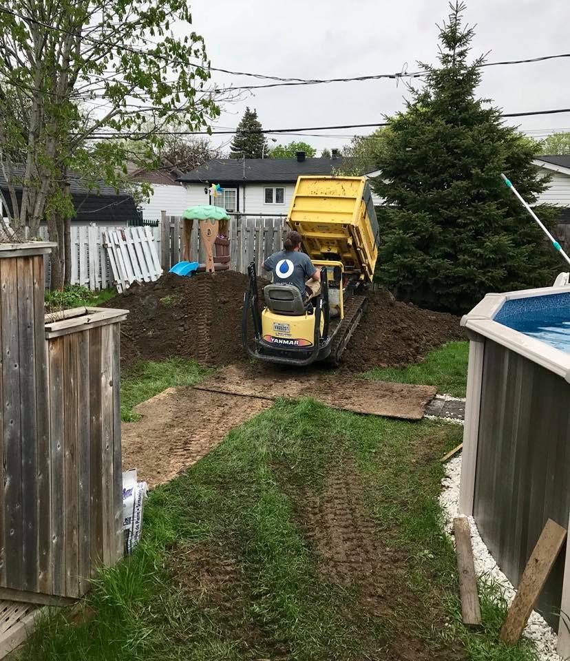 Une personne conduit un camion-benne jaune à chenilles, déchargeant de la terre dans un jardin près d'une piscine et d'une clôture blanche.