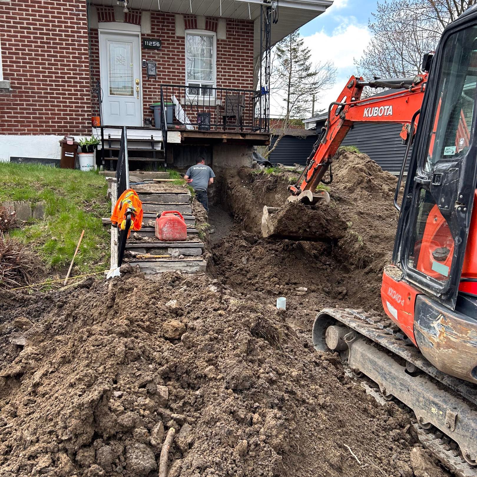 Une pelleteuse Kubota orange creuse une tranchée dans un jardin, à côté d'une maison en briques, près d'un escalier en bois.
