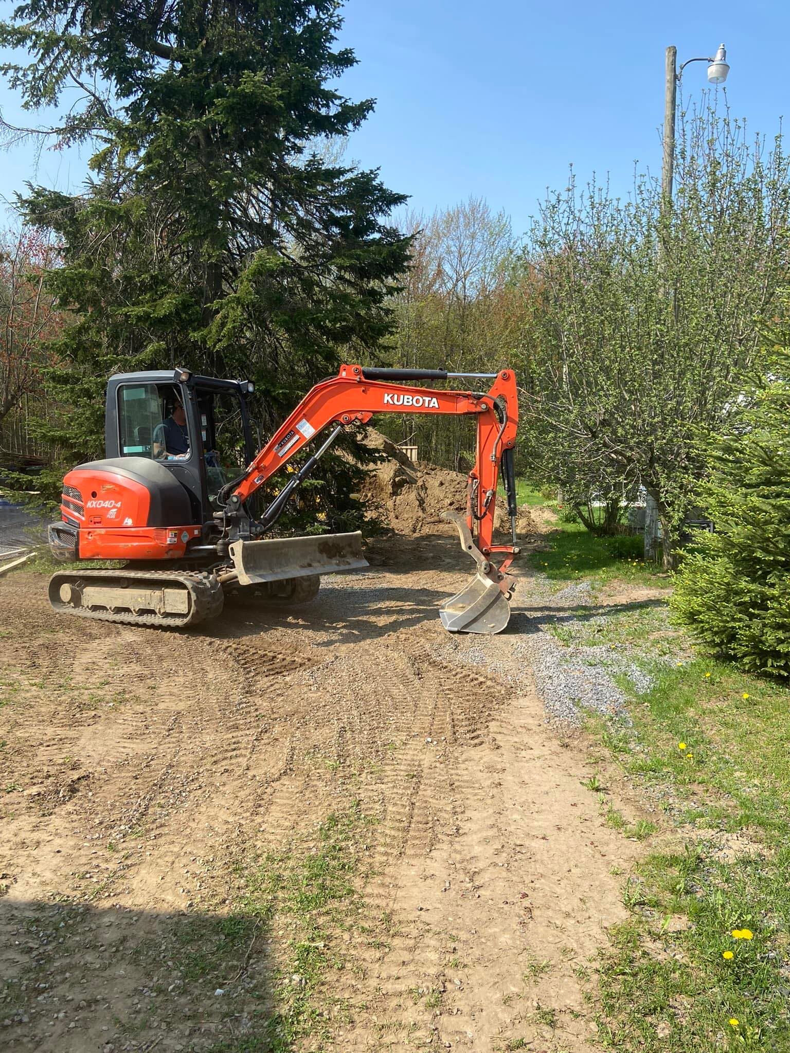 Une mini-pelle orange garée sur un chemin de terre dans un cadre boisé et herbeux par une journée ensoleillée.