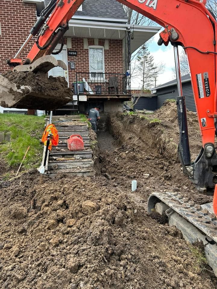 Une pelleteuse Kubota orange creuse une tranchée dans la cour d'une maison en briques tandis qu'un ouvrier se tient à proximité.