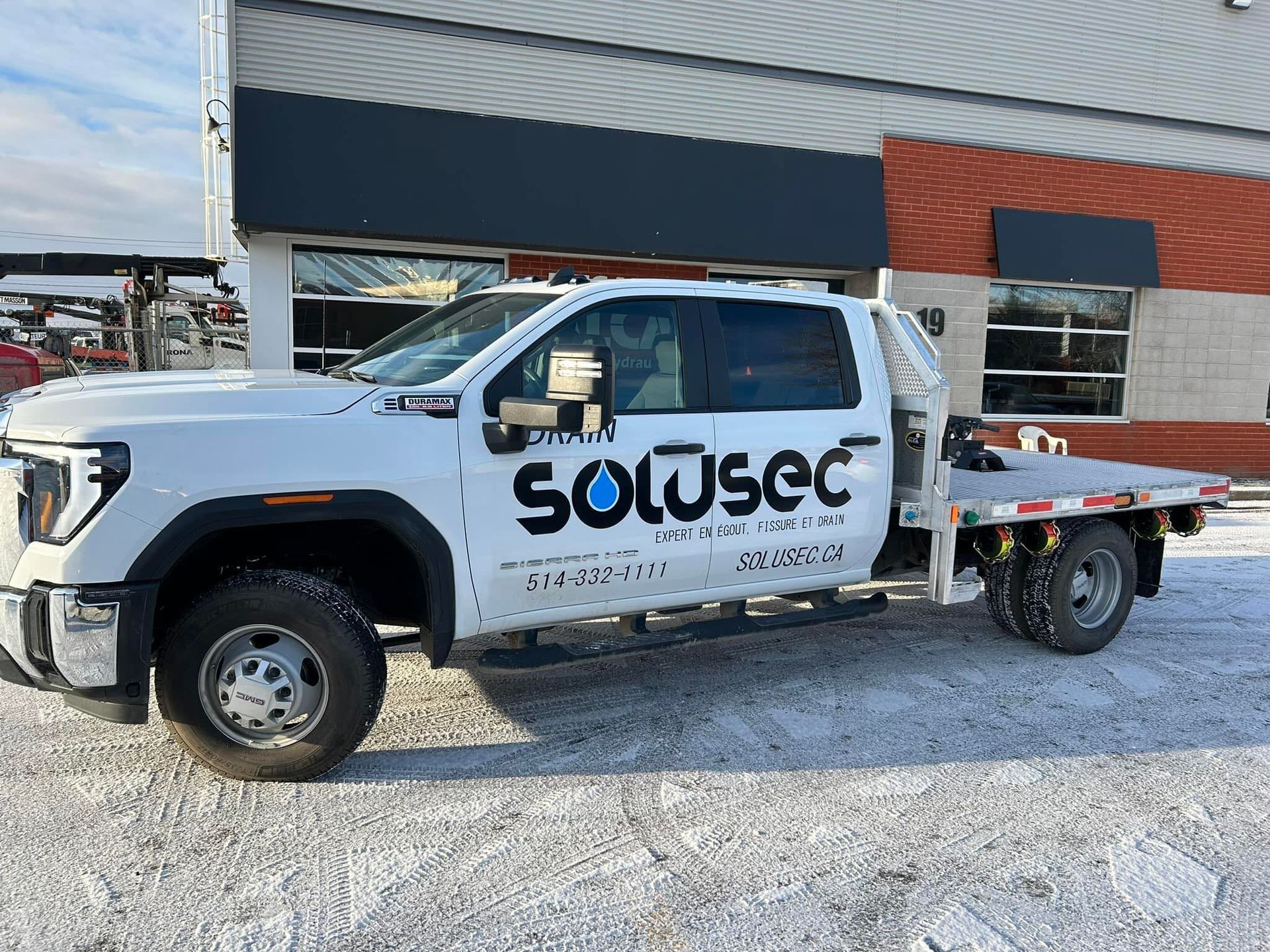 Un camion d'assistance Solusec blanc à plateau est stationné sur un terrain gravillonné devant un bâtiment.