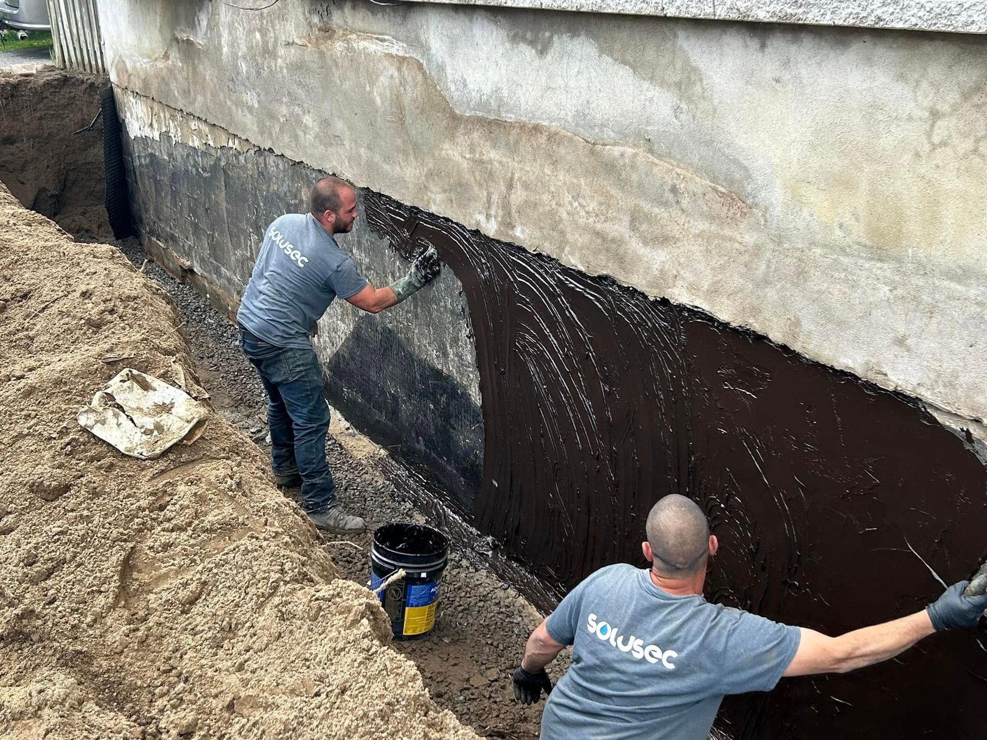 Deux ouvriers en chemises grises appliquent un revêtement imperméable noir sur un mur de fondation dans une tranchée.