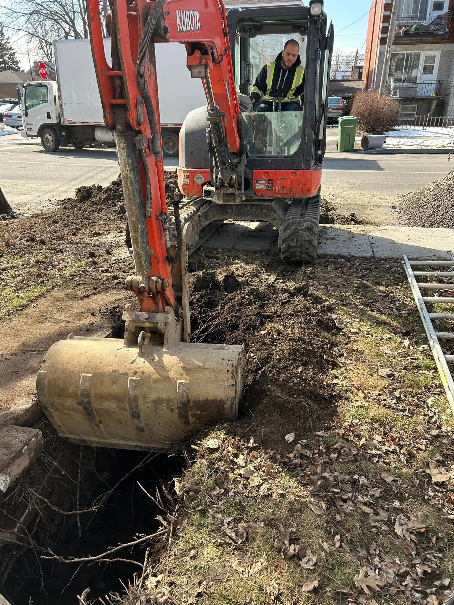 Une pelleteuse orange, avec un opérateur dans la cabine, est garée sur un chemin de terre à côté d'une tranchée remplie de feuilles mortes.