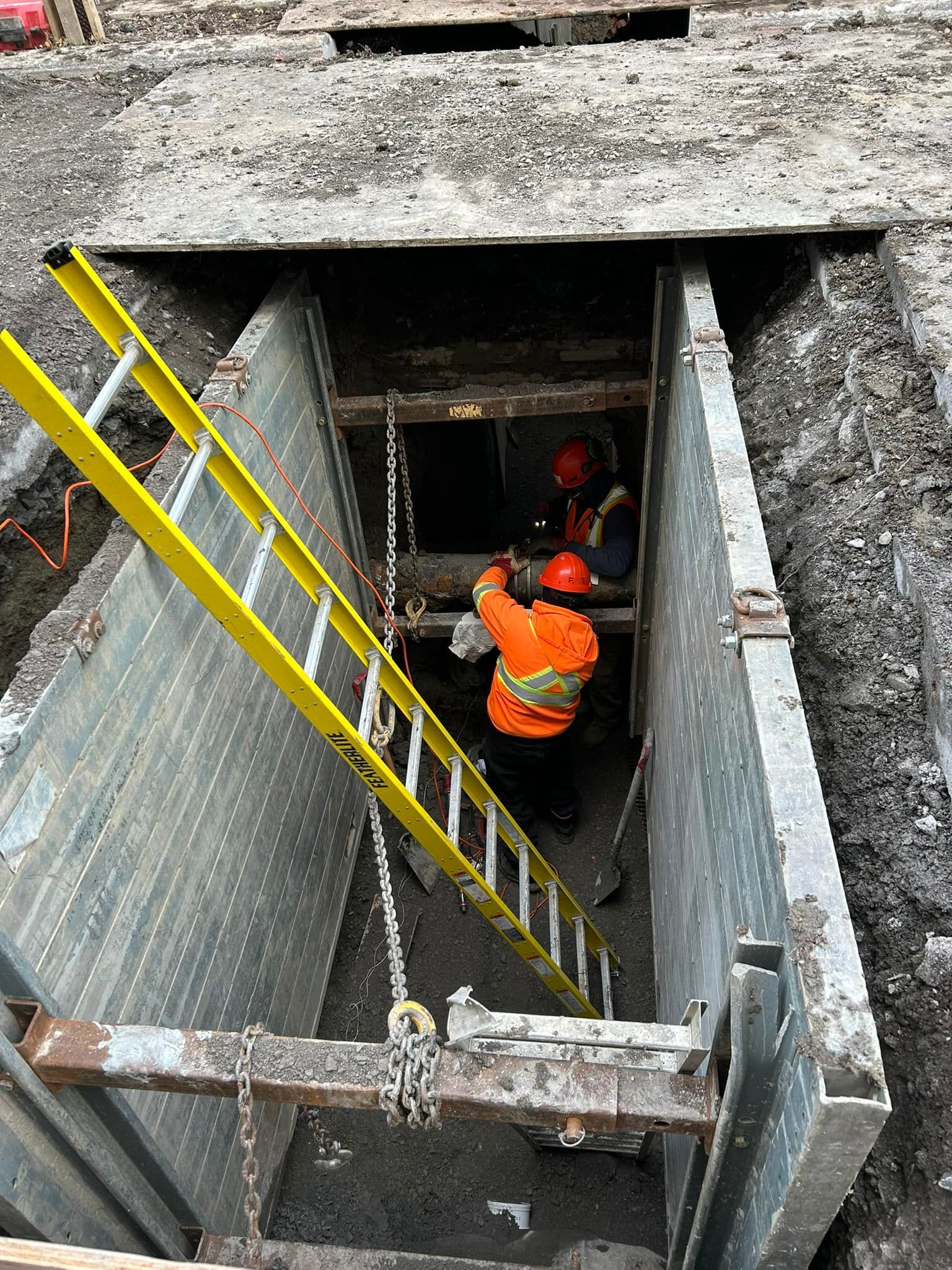 Deux ouvriers vêtus de gilets orange haute visibilité et de casques de chantier se trouvent à l'intérieur d'une tranchée d'excavation aux parois métalliques, à proximité d'une échelle.