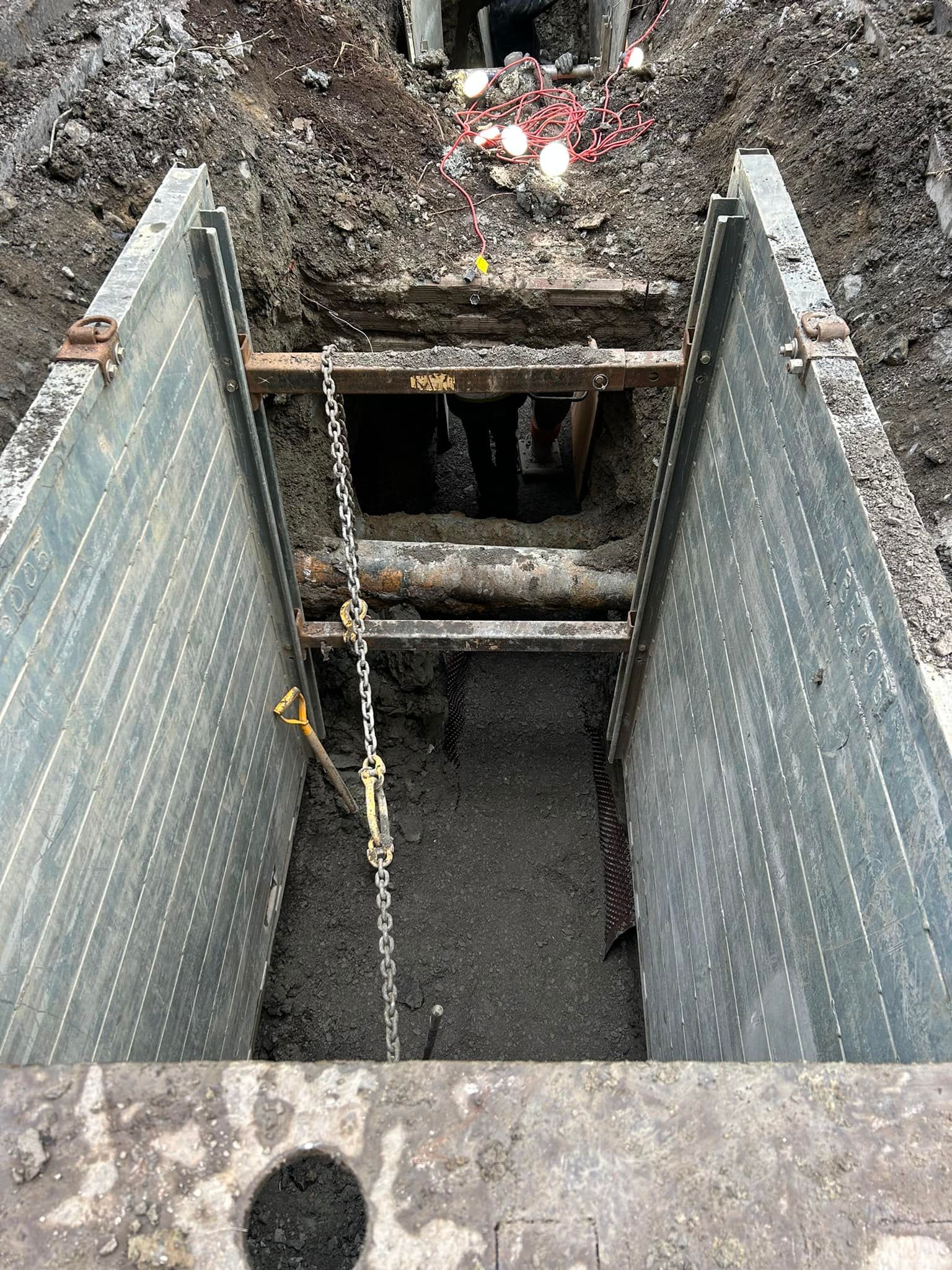 Vue de dessus d'une tranchée de construction ouverte avec des panneaux d'étaiement métalliques et une chaîne suspendue à l'intérieur.