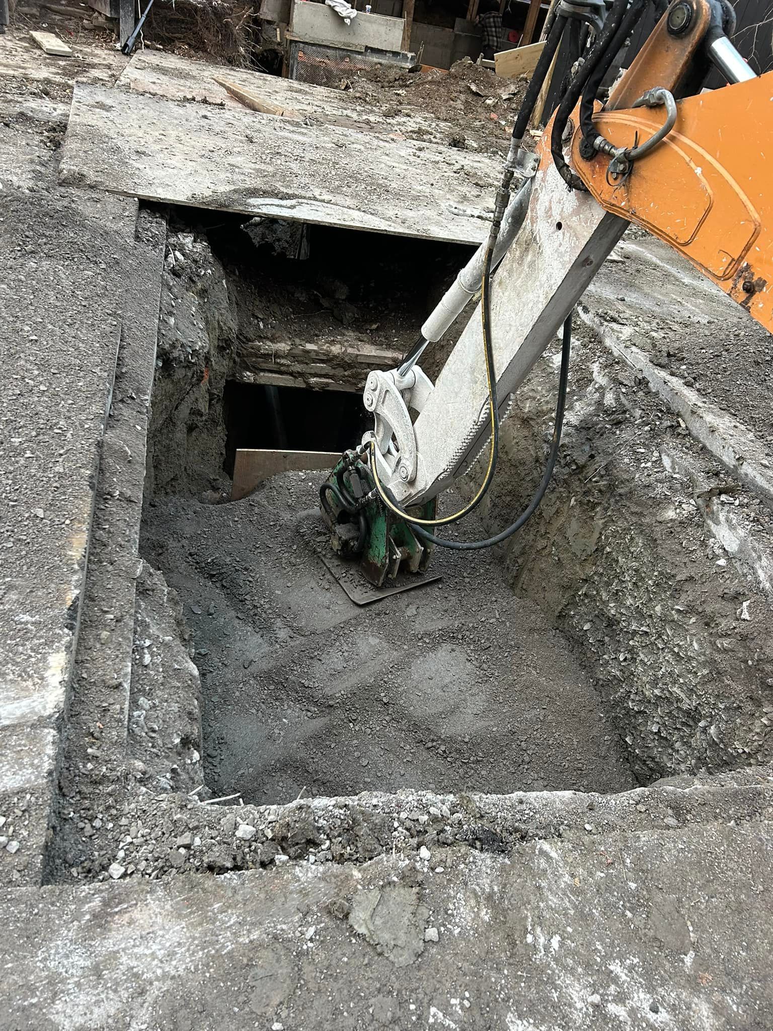 Un bras d'excavatrice orange utilise un outil de compactage hydraulique pour s'enfoncer dans un trou carré découpé dans une surface en béton.