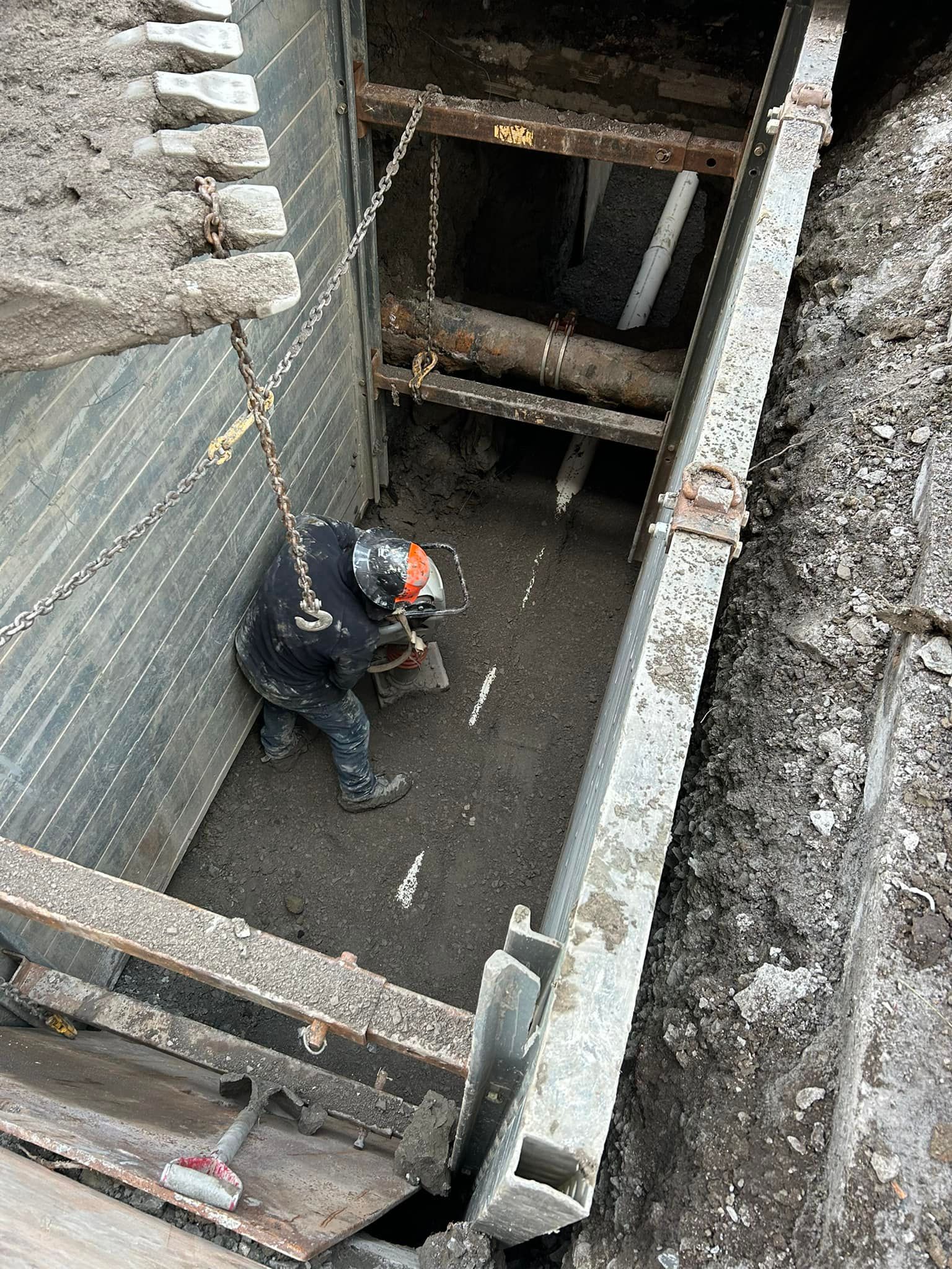 Un ouvrier portant un casque haute visibilité se tient à l'intérieur d'une profonde tranchée de construction étayée par des parois métalliques.