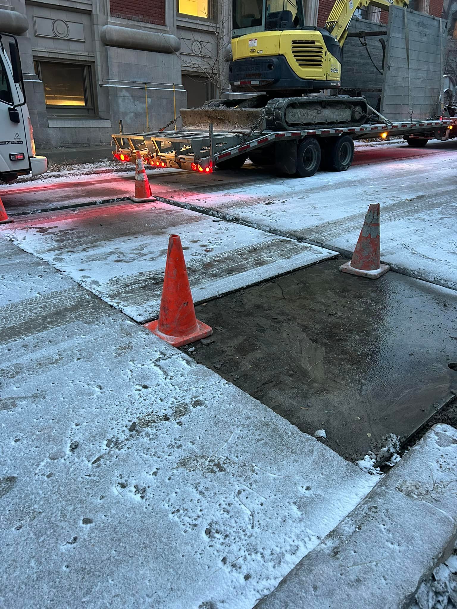 Des engins de chantier sur une remorque près d'un trou béant dans une rue enneigée, signalée par des cônes de signalisation orange.