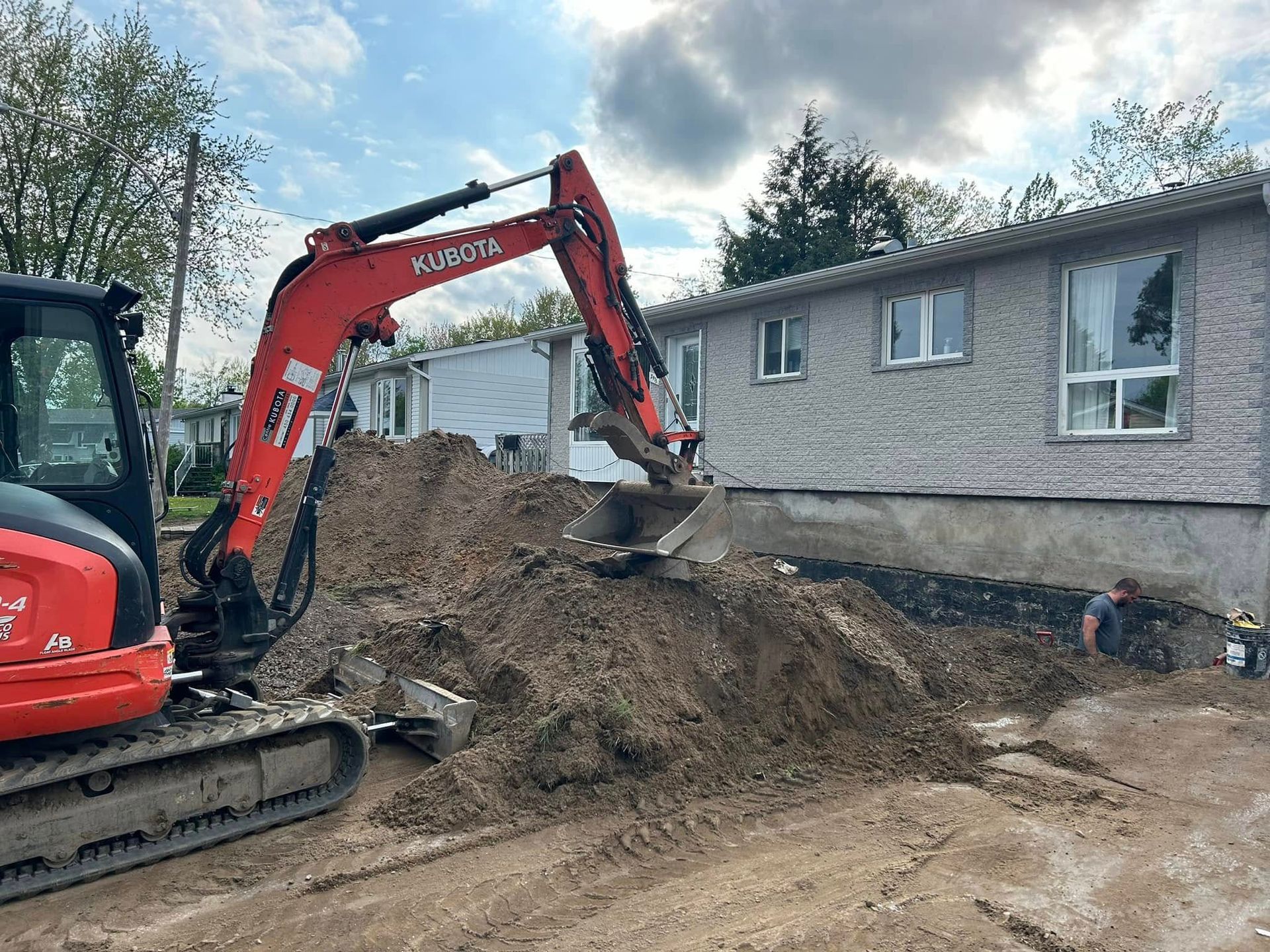 Une pelleteuse orange creuse une tranchée le long d'une maison de couleur claire, tandis qu'un ouvrier travaille près des fondations.
