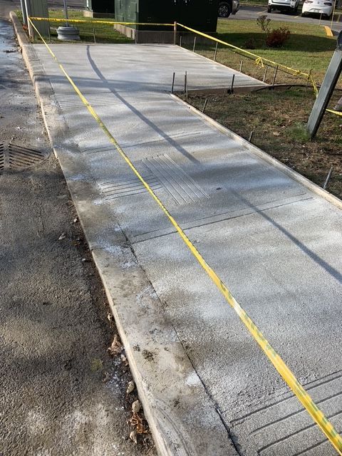 Une portion de trottoir en béton fraîchement coulé, délimitée par un ruban de signalisation jaune, dans un espace extérieur herbeux.