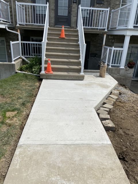 Un trottoir en béton fraîchement coulé mène à un escalier avec des rampes blanches et deux cônes de signalisation orange.