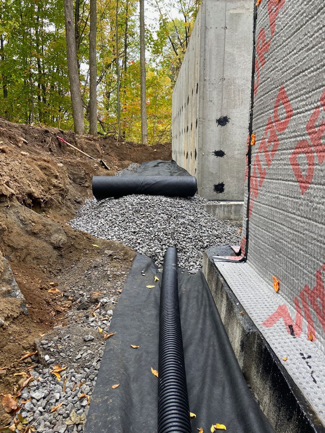 Un tuyau de drainage noir repose sur une toile de paillage dans une tranchée à côté d'un mur de fondation rempli de gravier et d'un rouleau de toile.