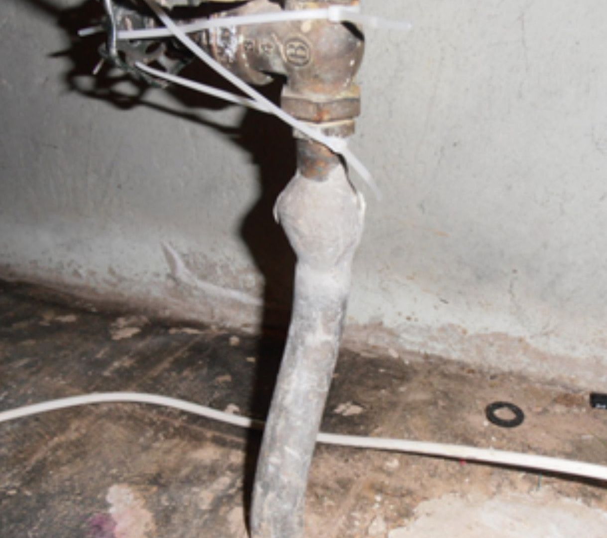 Un tuyau de plomberie en métal avec un joint corrodé et bombé, relié à une vanne et fixé par des colliers de serrage en plastique blanc.