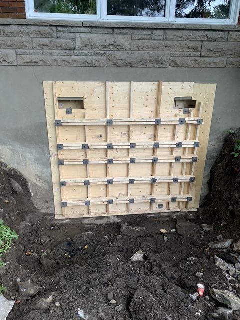 Une structure de coffrage en contreplaqué est adossée à un mur extérieur de sous-sol sur un chantier de construction, la terre étant excavée en dessous.