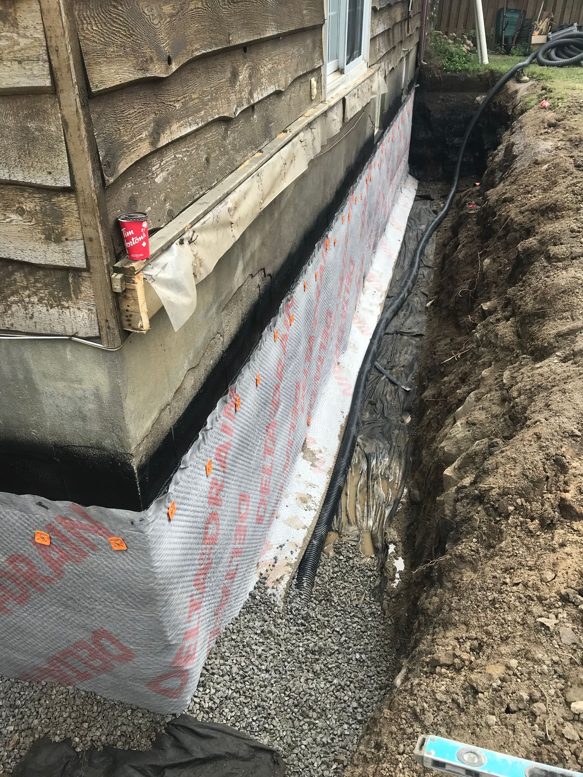 Un mur de fondation de maison avec une nouvelle membrane de drainage étanche installée, à côté d'une tranchée remplie de gravier.