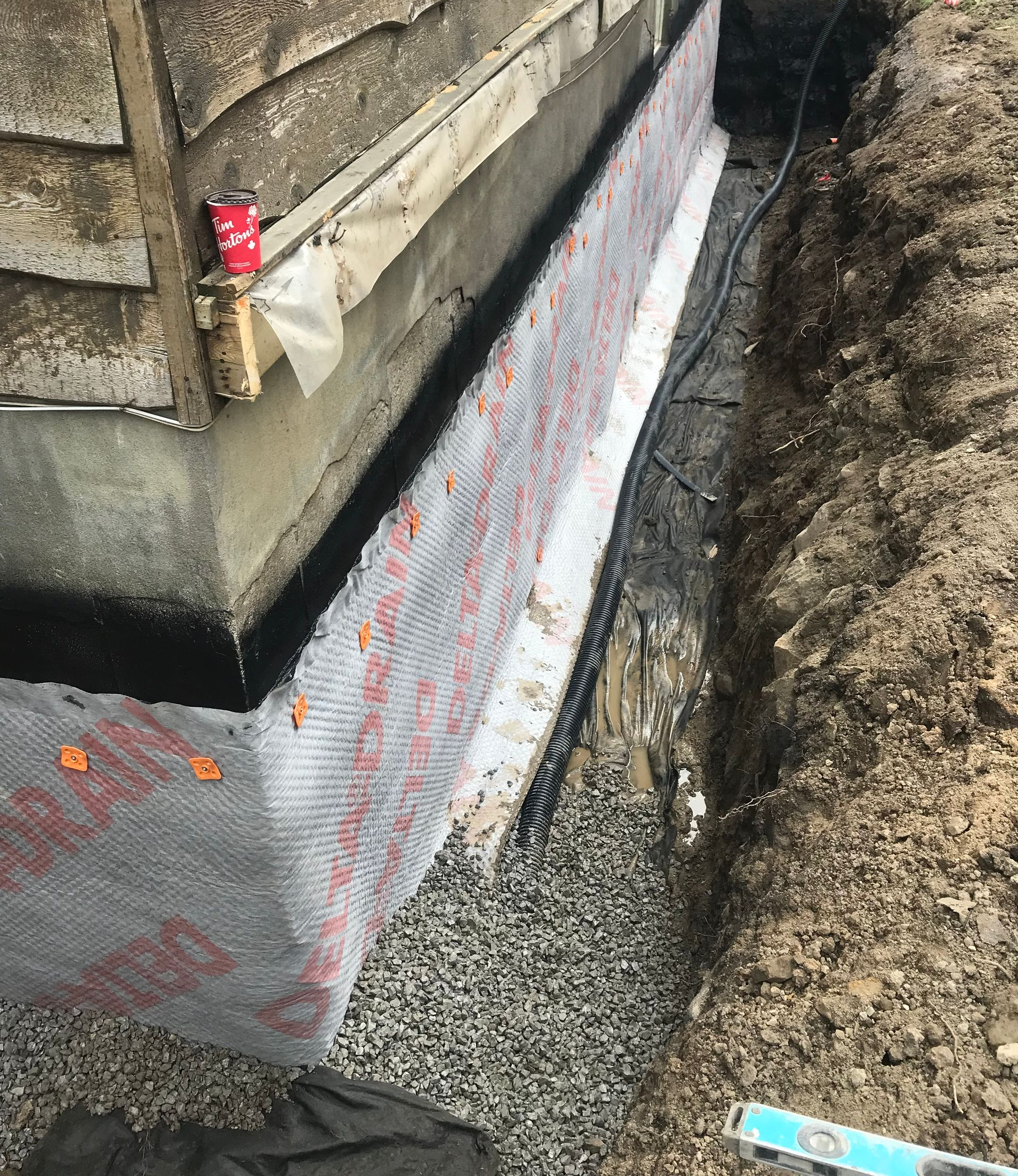 Une tranchée de fondation montrant un mur en béton protégé par une membrane de drainage alvéolée et une base de gravier.