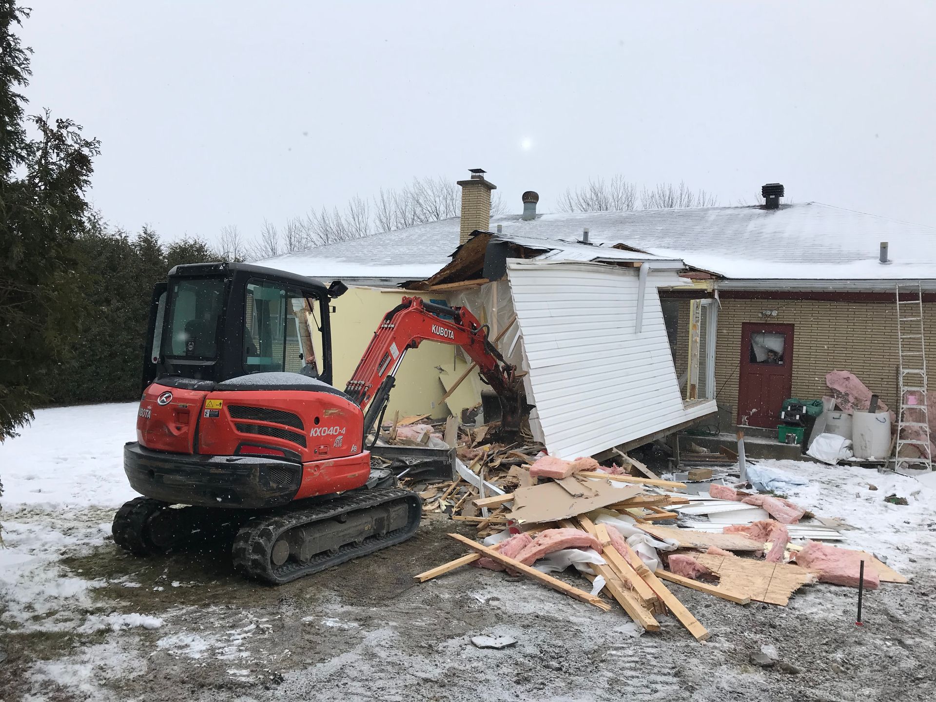 Une pelleteuse rouge démolit le mur d'une maison en hiver, laissant des débris et de l'isolant éparpillés sur le sol enneigé.