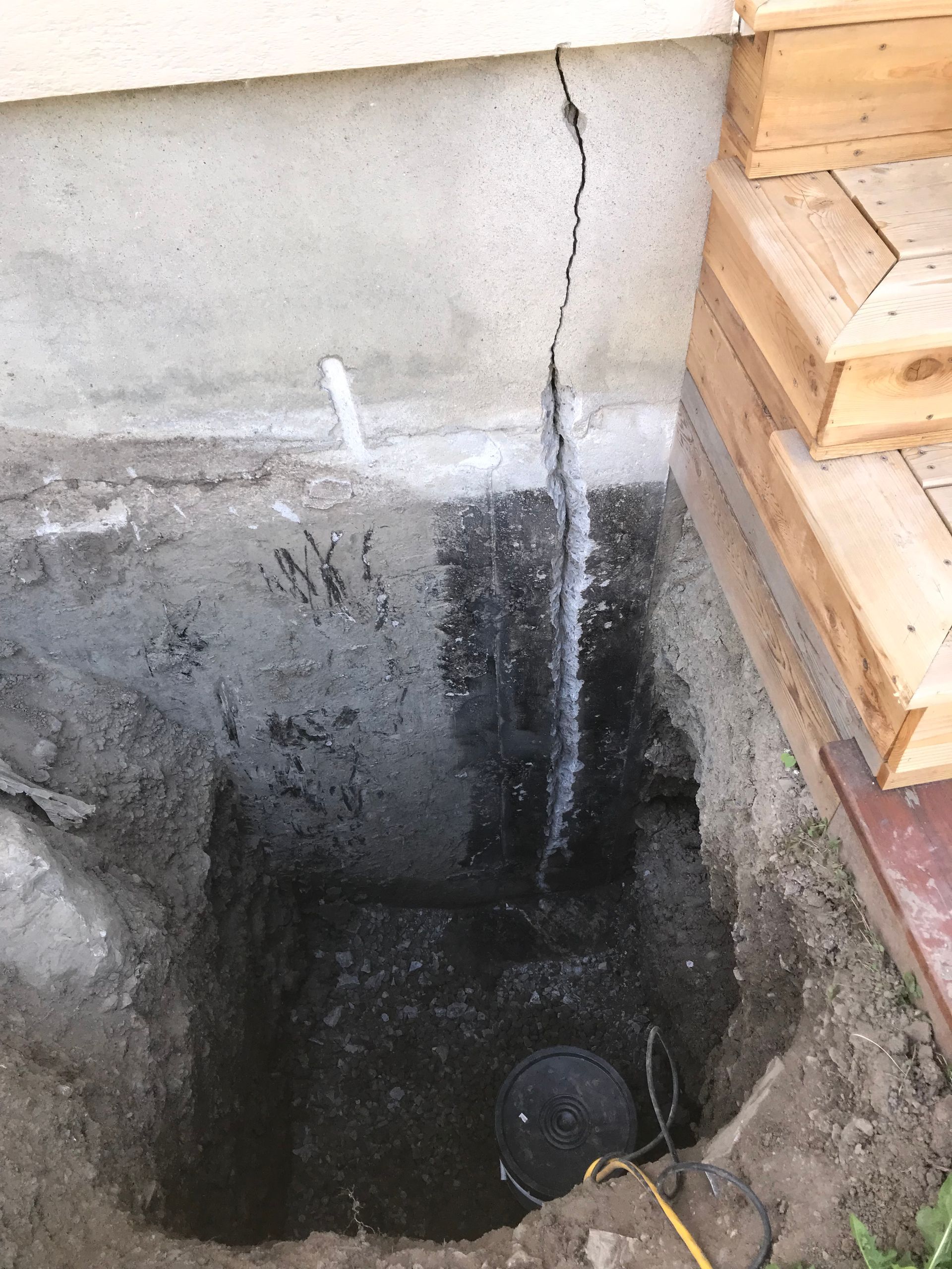 Une fissure verticale dans un mur de fondation en béton gris, mise au jour par une fosse creusée à côté des marches d'un porche en bois.